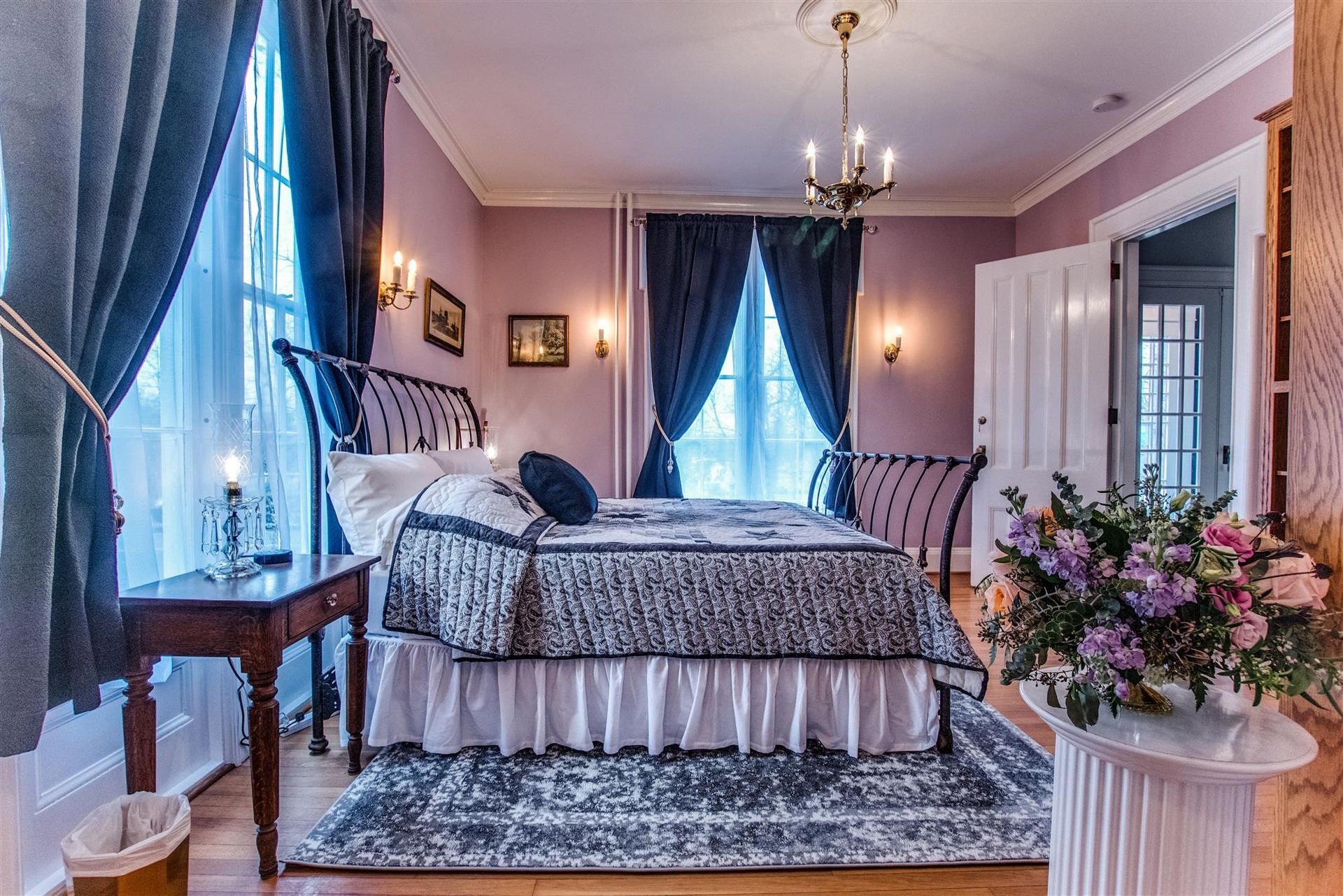 Elegant lavender bedroom with a canopy bed, blue curtains, chandelier, and flower arrangement