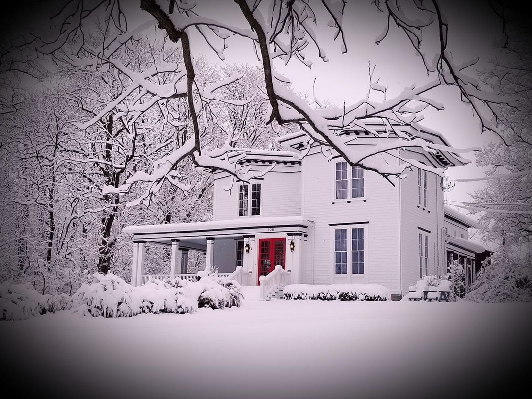 Snow-covered white house with a red front door and bare trees in winter