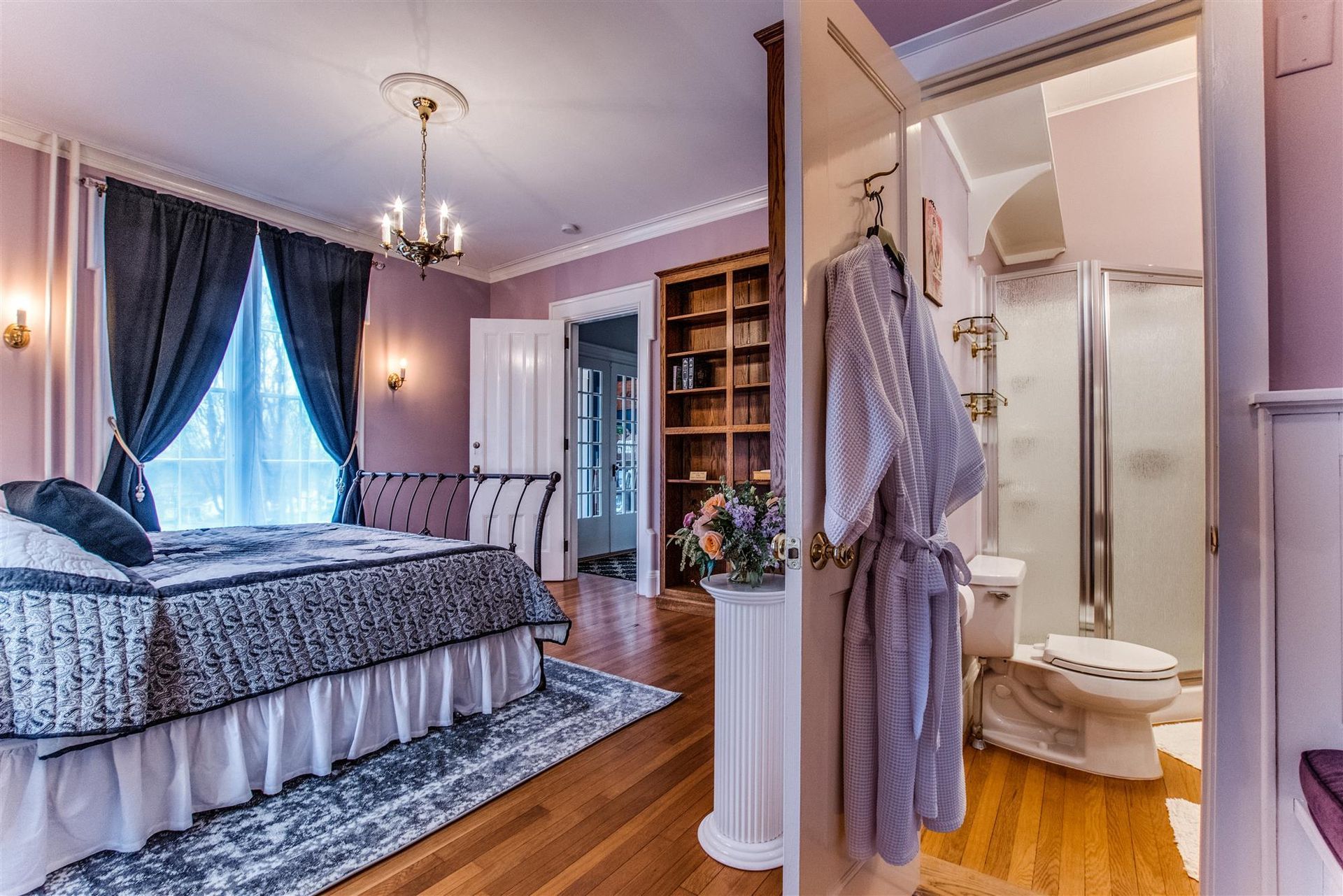 Bright bedroom with purple drapes, bed, desk, and a bathroom vanity visible through an open doorway