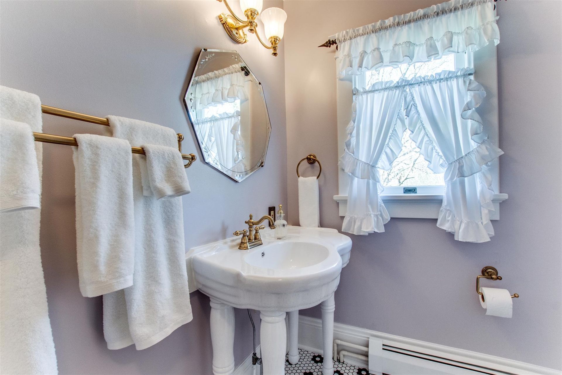 White bathroom with pedestal sink, wall mirror, brass fixtures, and lace-curtained window