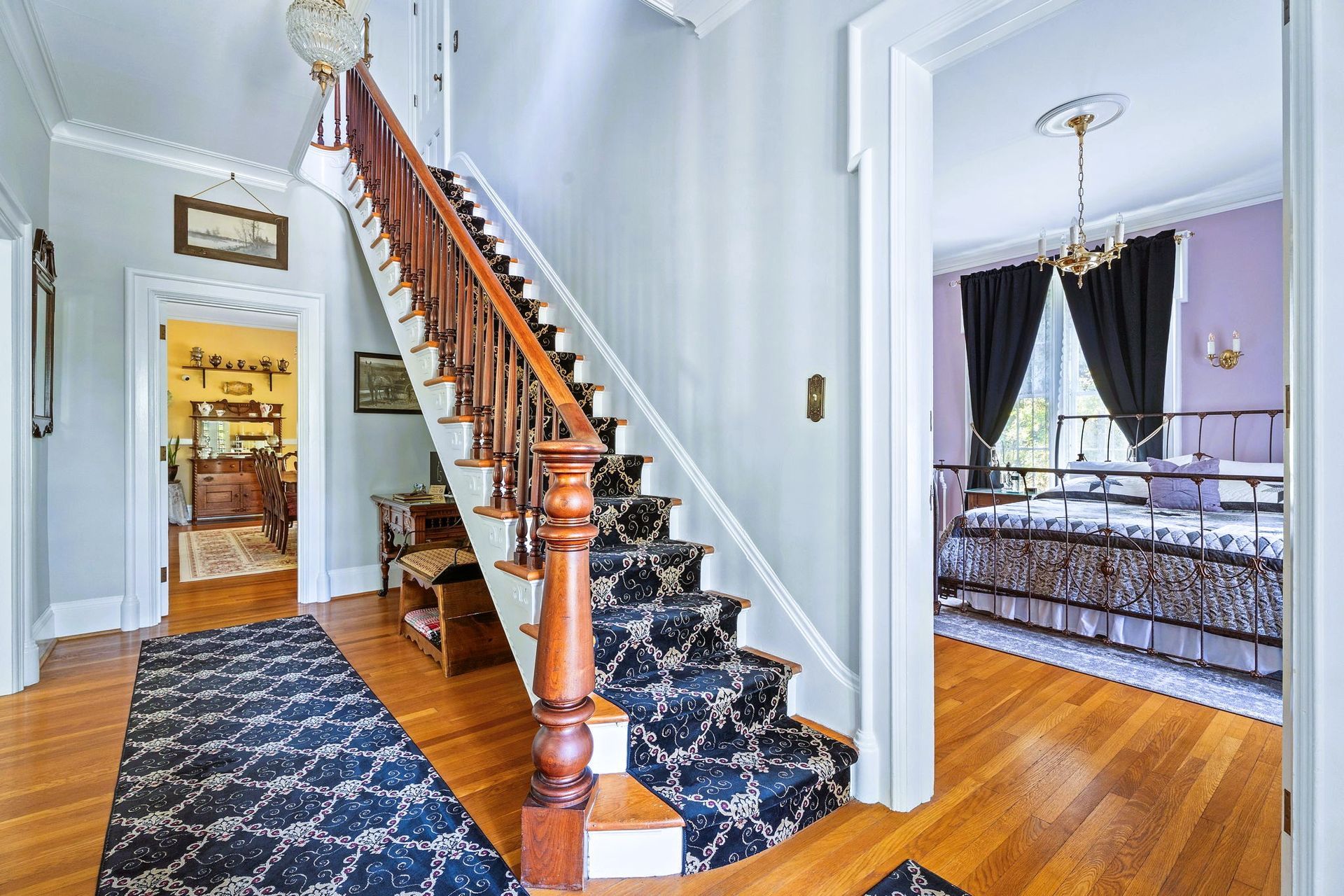 Hallway with ornate wooden staircase, blue patterned runner, and doorway to a purple bedroom