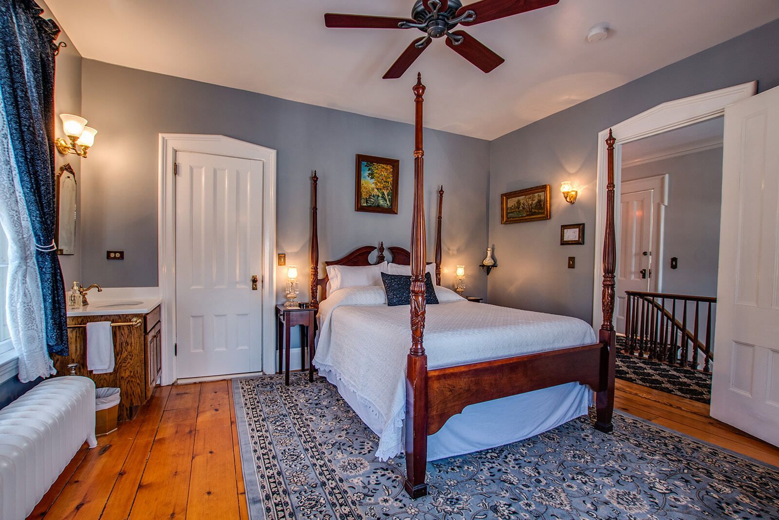 Bedroom with four-poster bed, ceiling fan, patterned rug, and gray walls with framed art.