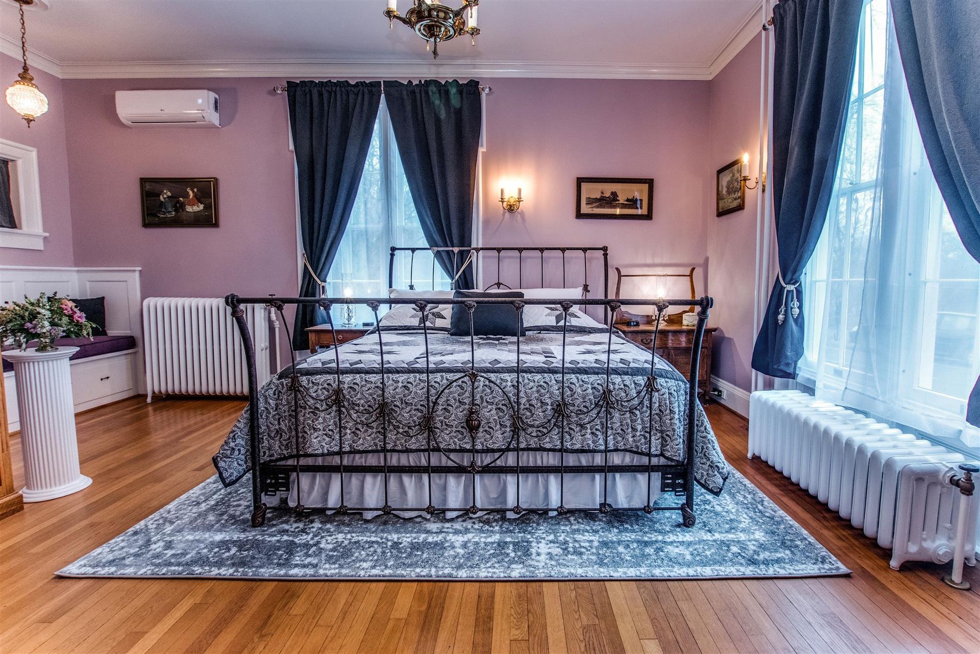 Lavender bedroom with a black wrought-iron bed, hardwood floors, and large blue-curtained windows.
