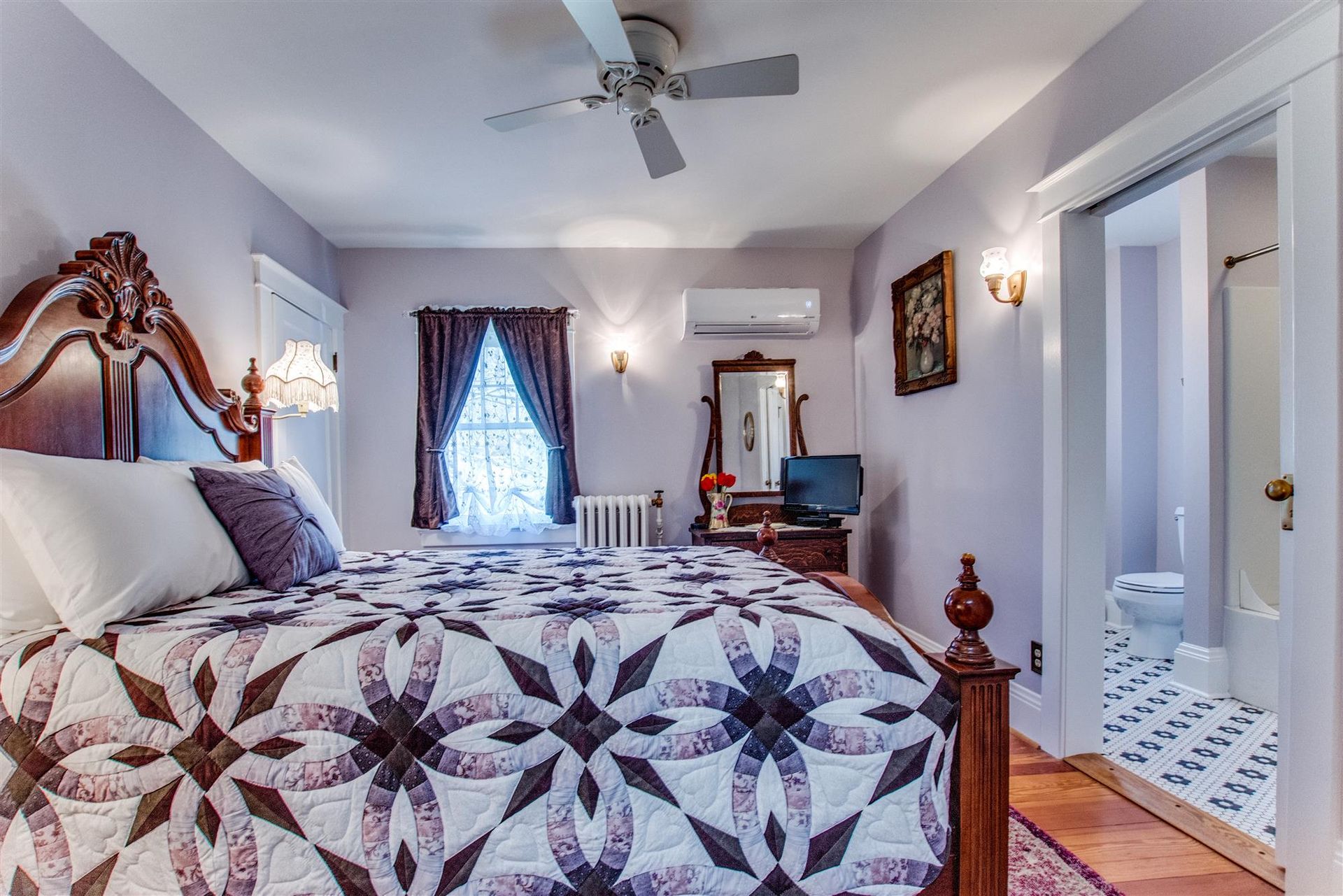 Bedroom with quilted bed, white walls, ceiling fan, window curtains, and adjoining bathroom visible
