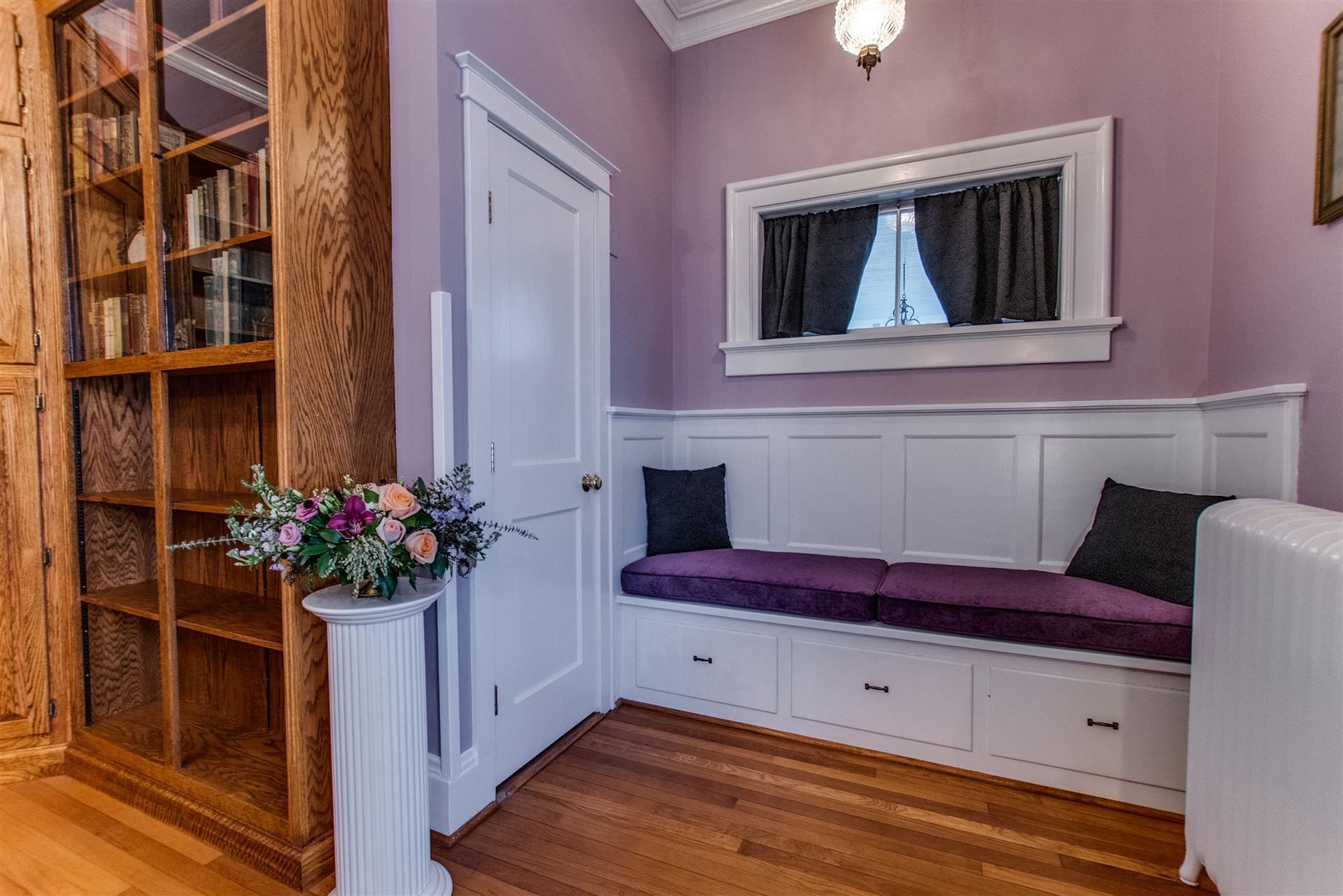 Cozy window bench nook with purple cushion, built-in drawers, and a vase of flowers in a wood-floored room