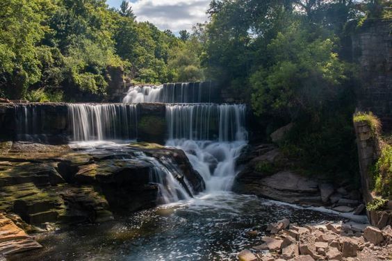 Wide waterfall cascading over rocky ledges in a lush green forest under a partly cloudy sky