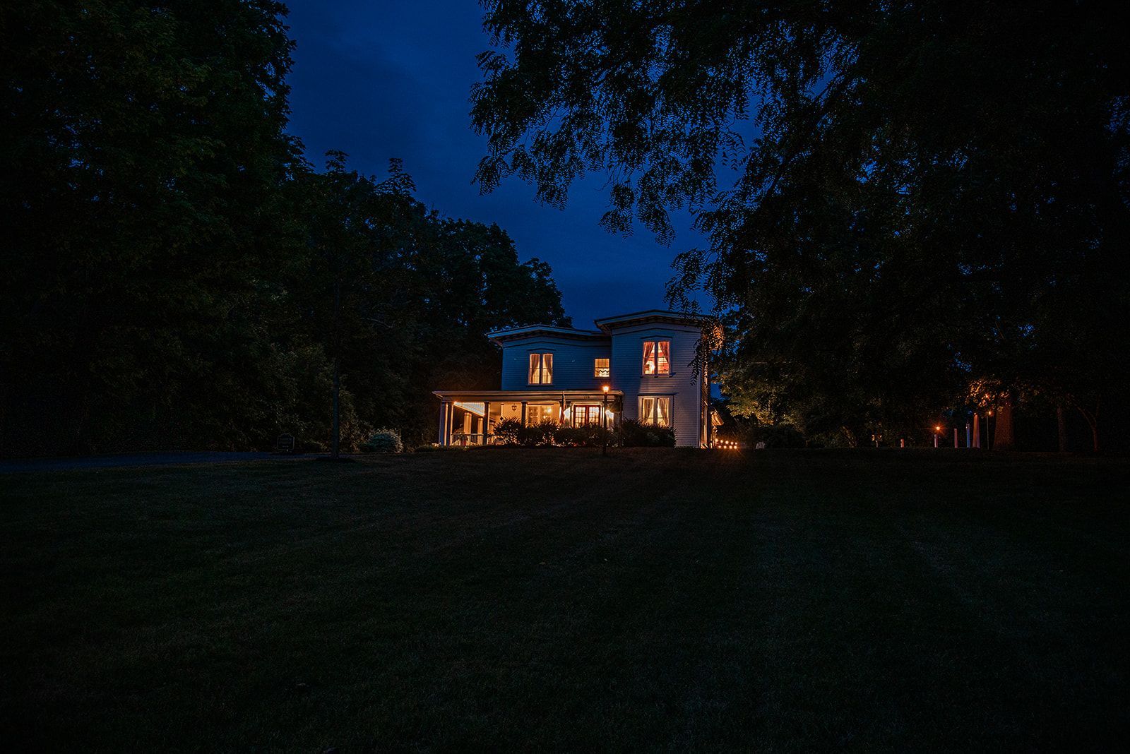 A lit house at night with warm lights glowing through windows, surrounded by dark trees and lawn.