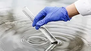 A gloved hand dips a clear glass test tube into rippling, murky water.