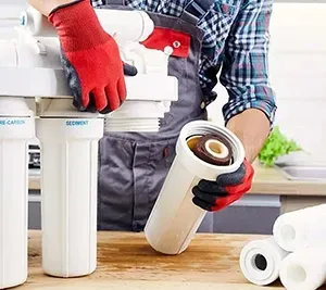 A technician in work clothes and red gloves replaces a filter cartridge in a multi-stage water filtration system.