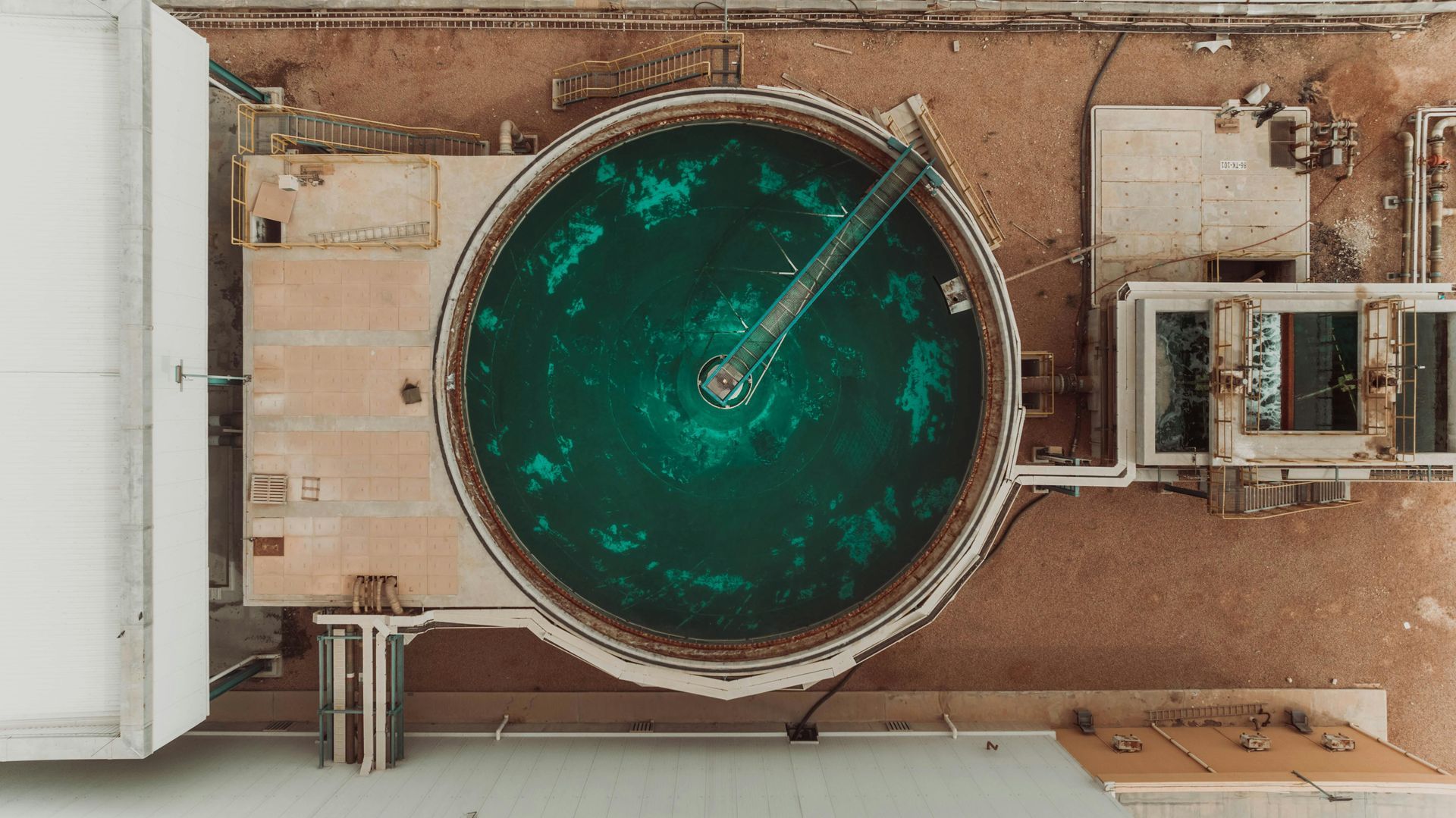 An aerial view of a circular, turquoise water treatment tank surrounded by concrete structures and sandy ground.
