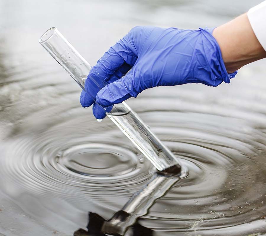 A gloved hand dips a clear glass test tube into a body of water to collect a sample.