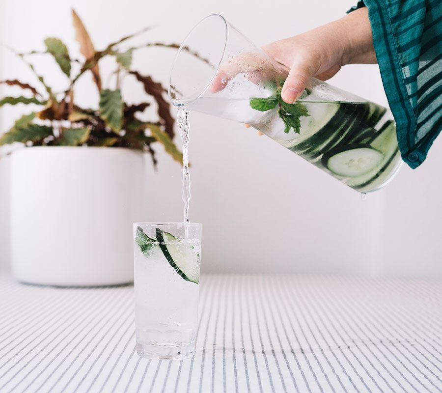 A hand pours cucumber and mint-infused water from a clear pitcher into a glass, with a potted plant in the background.
