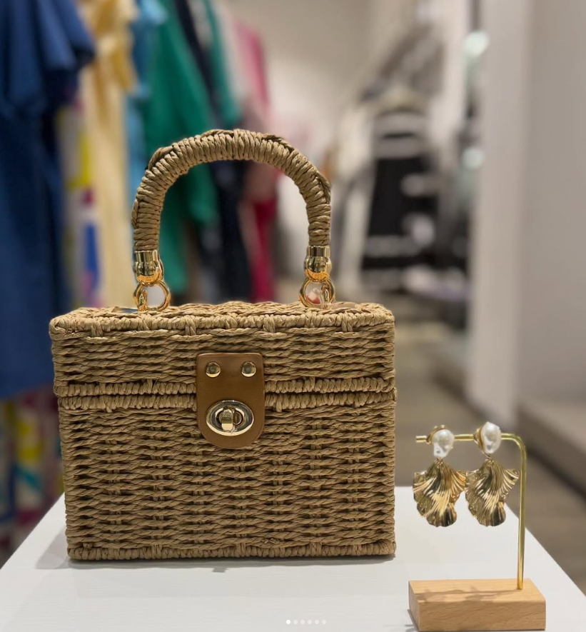 Wicker handbag with gold accents and earrings on a stand displayed on a white table in a boutique.