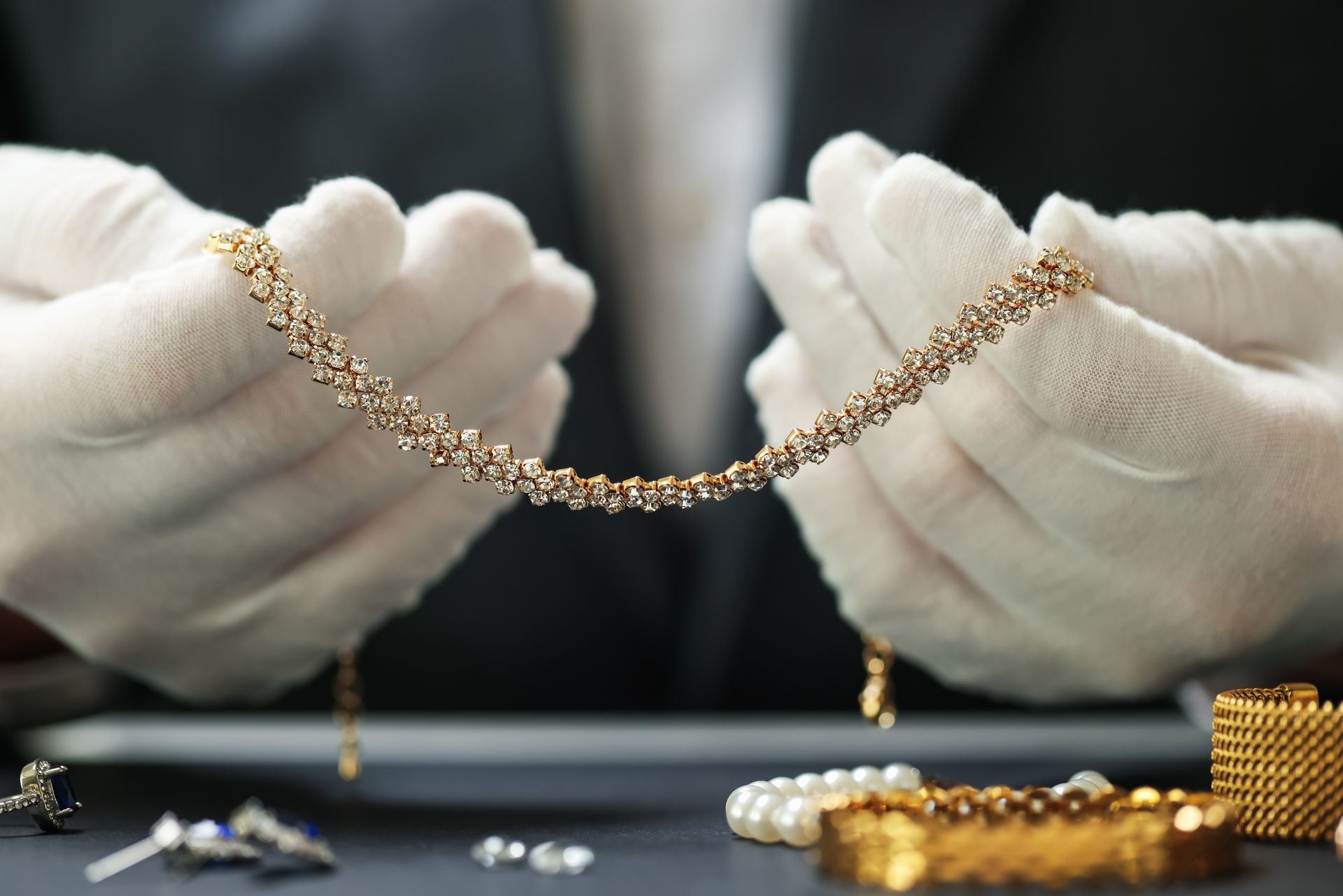 Hands in white gloves hold a gold necklace with sparkling gems; jewelry displayed on a dark surface.