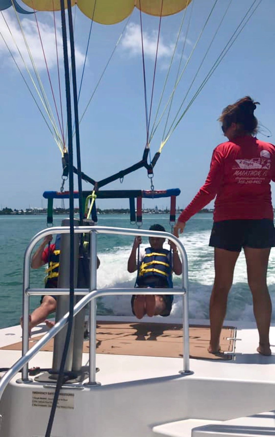A woman in a red shirt is standing on a boat with people parasailing