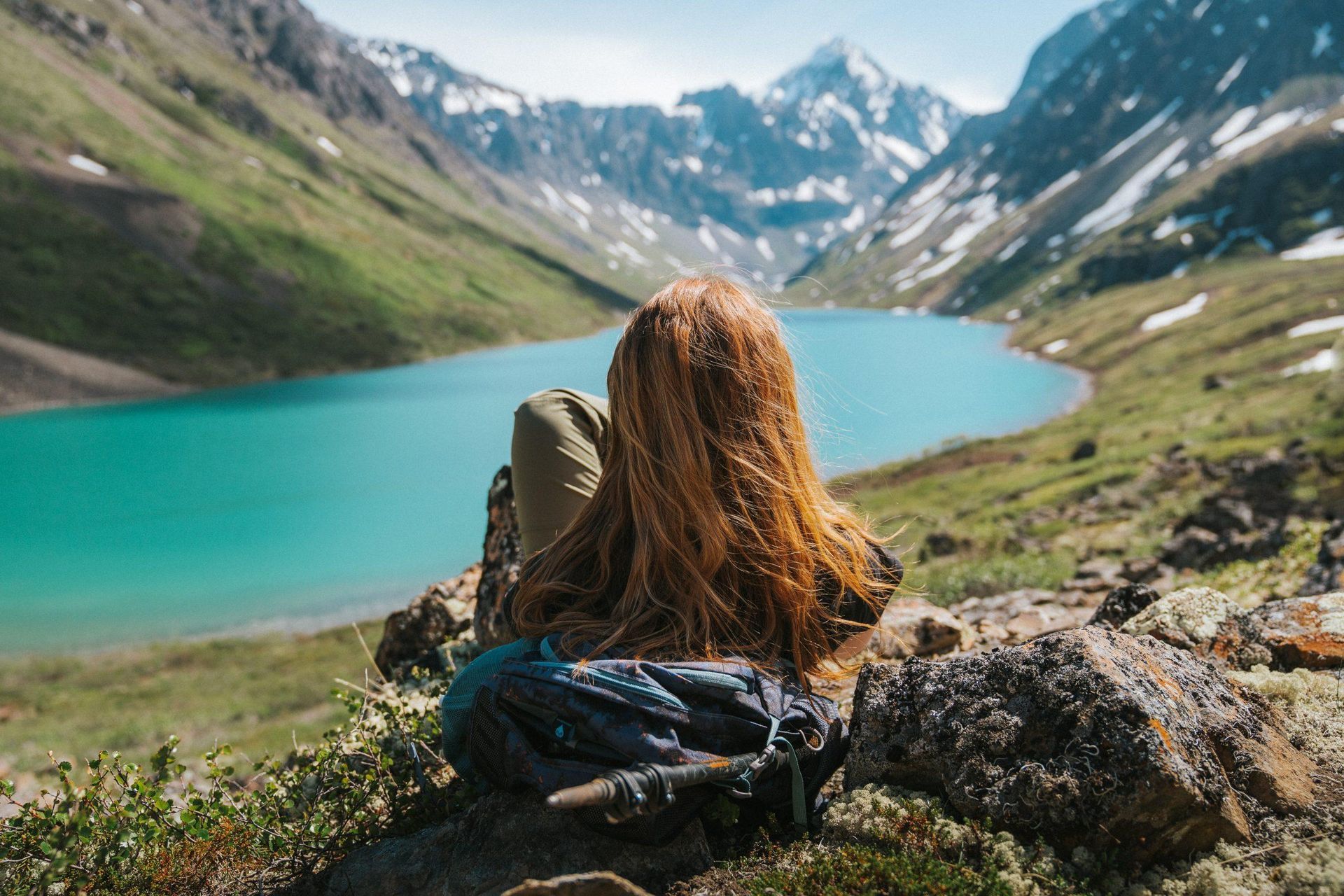 Woman with long red hair sits on a rock, gazing at a turquoise lake surrounded by mountains.