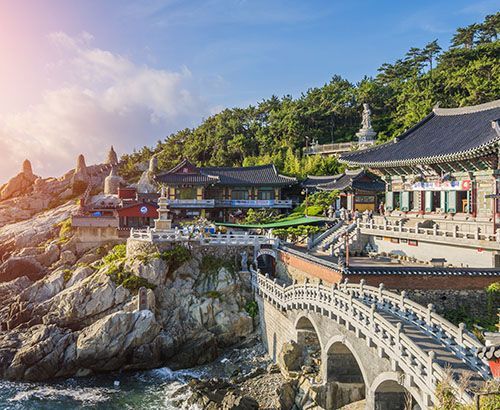A temple complex perched on a rocky coastline, with a bridge, statues, and buildings under a sunny sky.