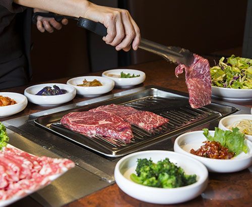 Person grilling meat at a table with side dishes, hands holding tongs, and a close-up of raw meat.