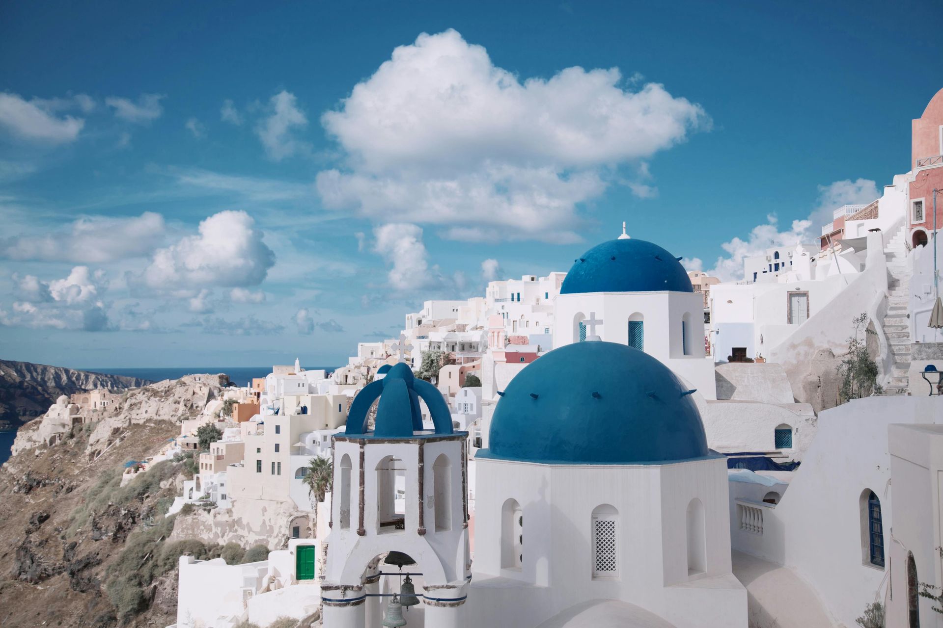 Whitewashed buildings with blue domes on a cliff in Santorini, Greece, under a bright blue sky with clouds.