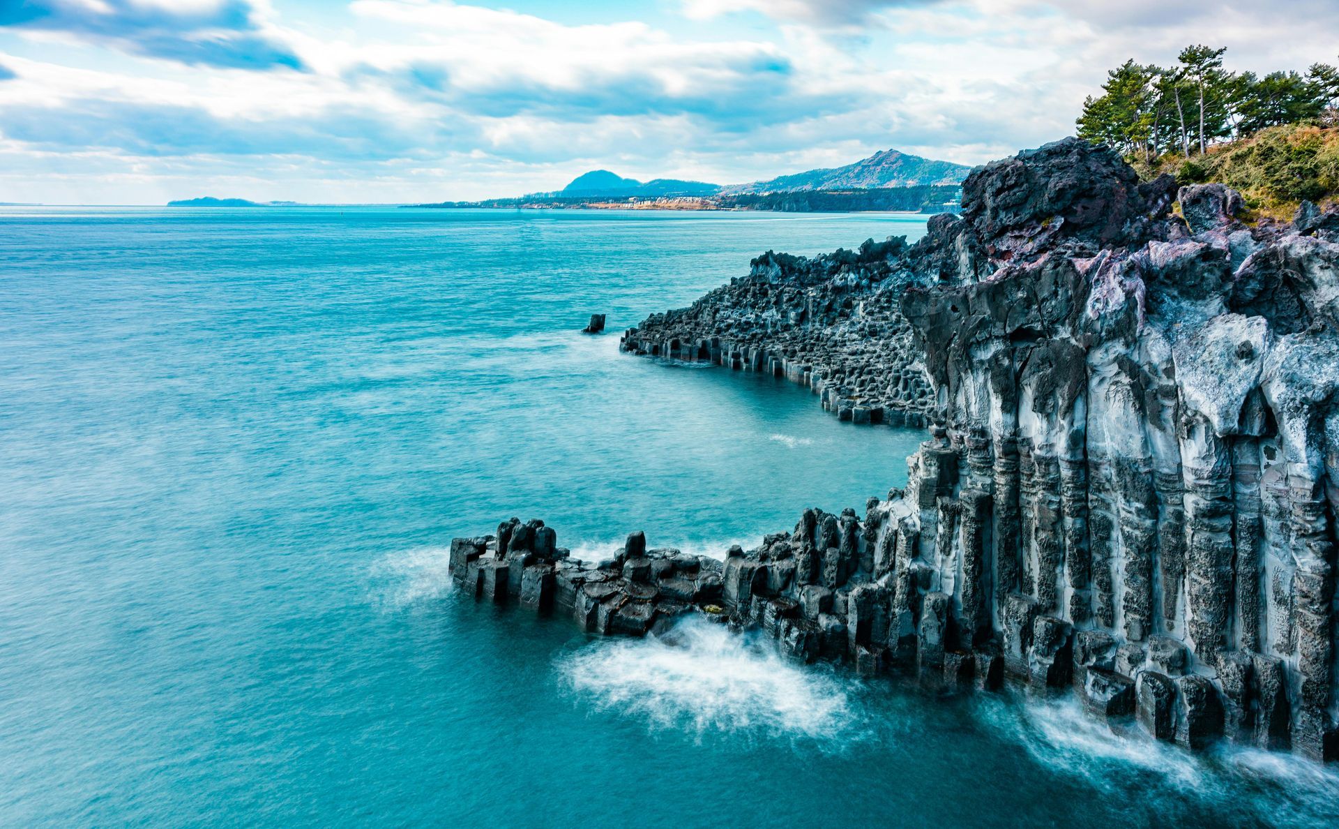 Ocean waves crashing against a rugged, dark rock cliff under a cloudy sky.