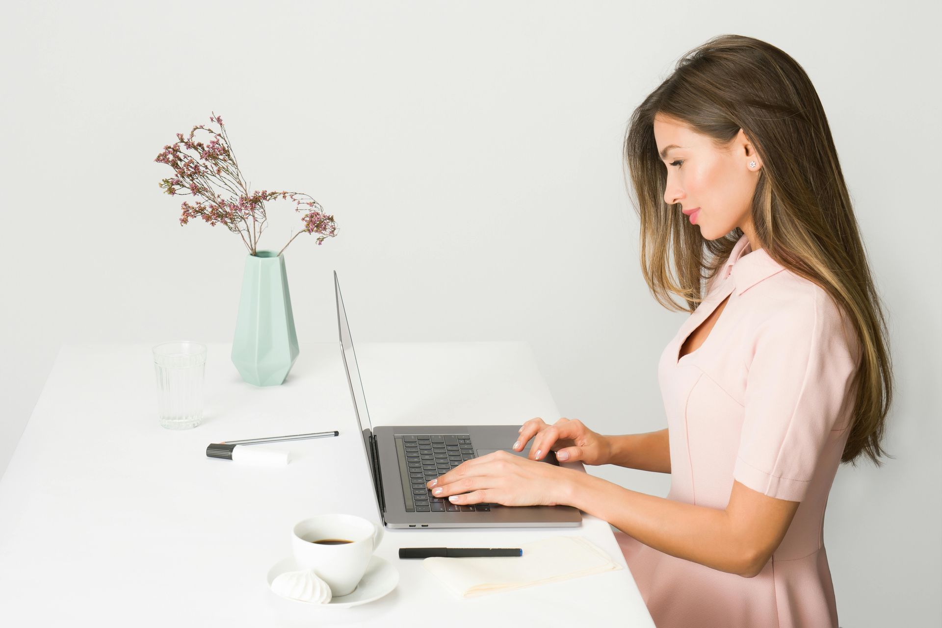 Woman typing on laptop at white desk, coffee, vase of flowers.