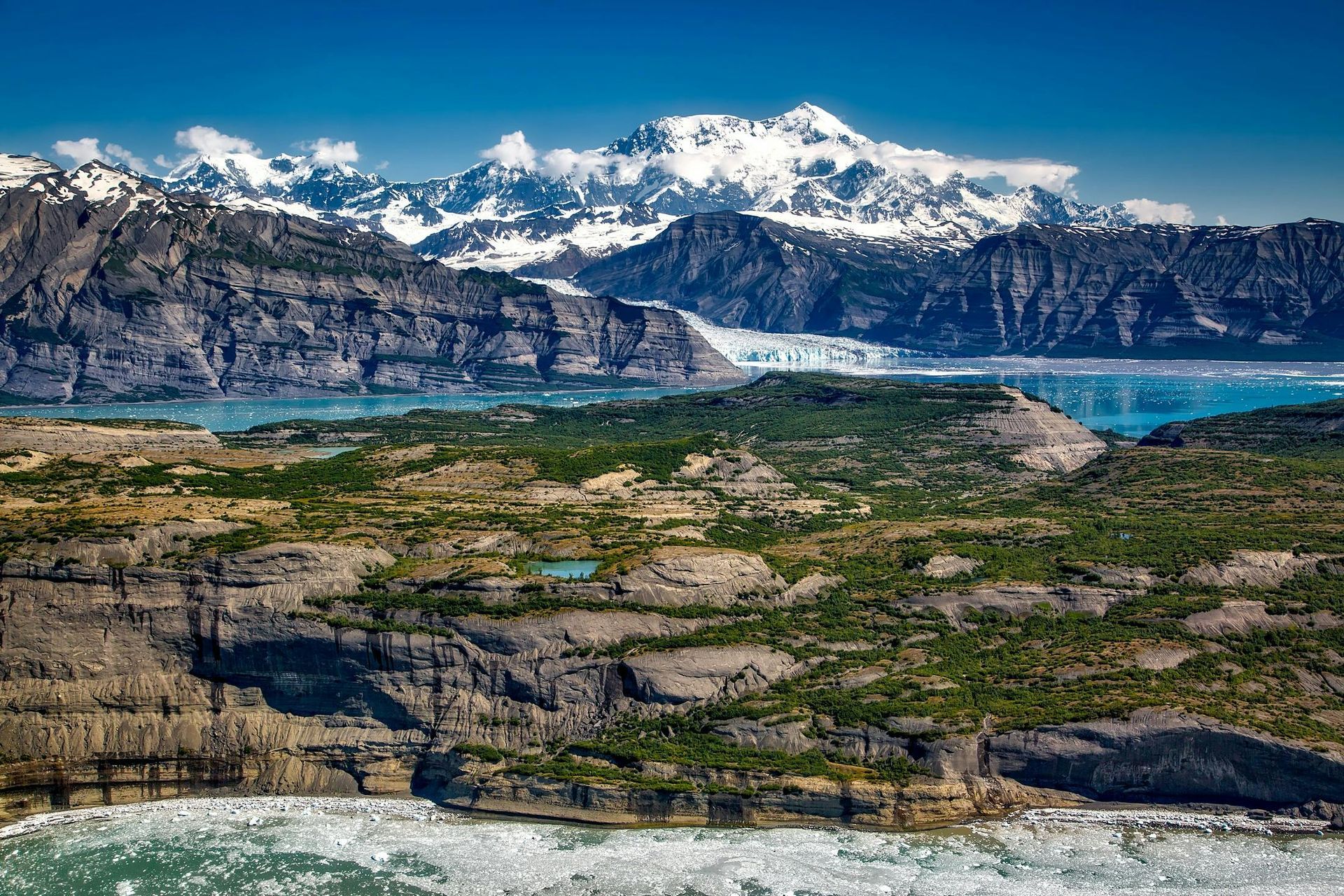 Rocky landscape with mountains, glacier, and water under a blue sky.