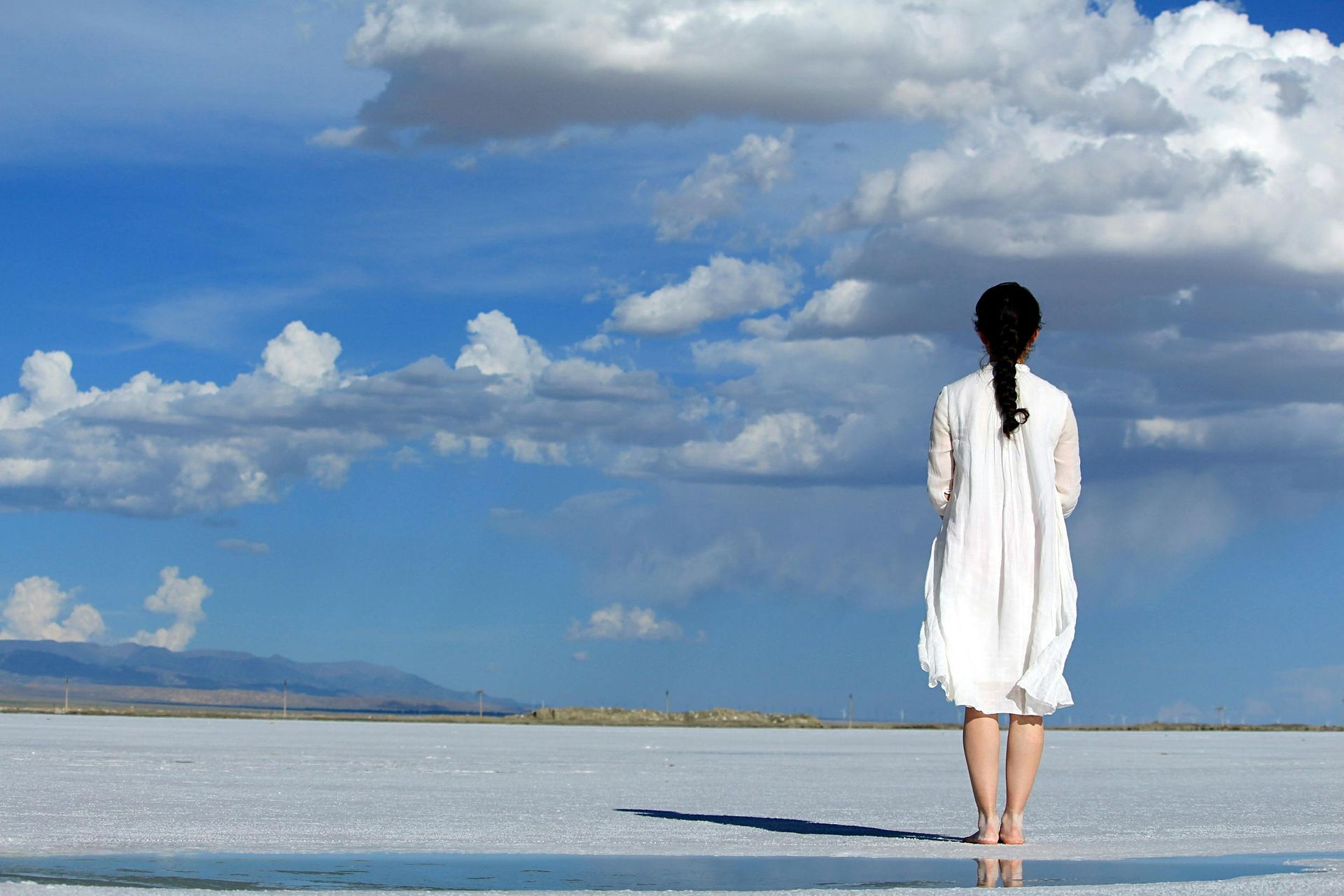 Woman in white dress standing on reflective water, looking at blue sky with clouds.
