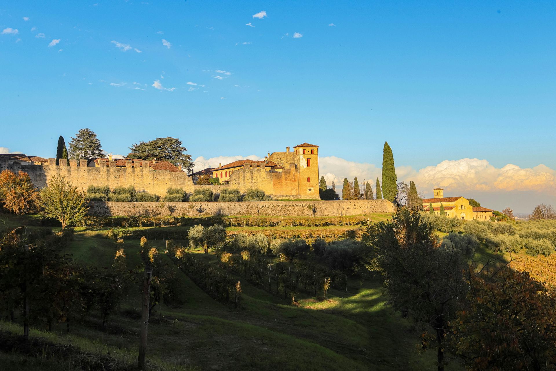 Stone wall and buildings atop a green hillside under a blue sky, possibly Italian countryside.