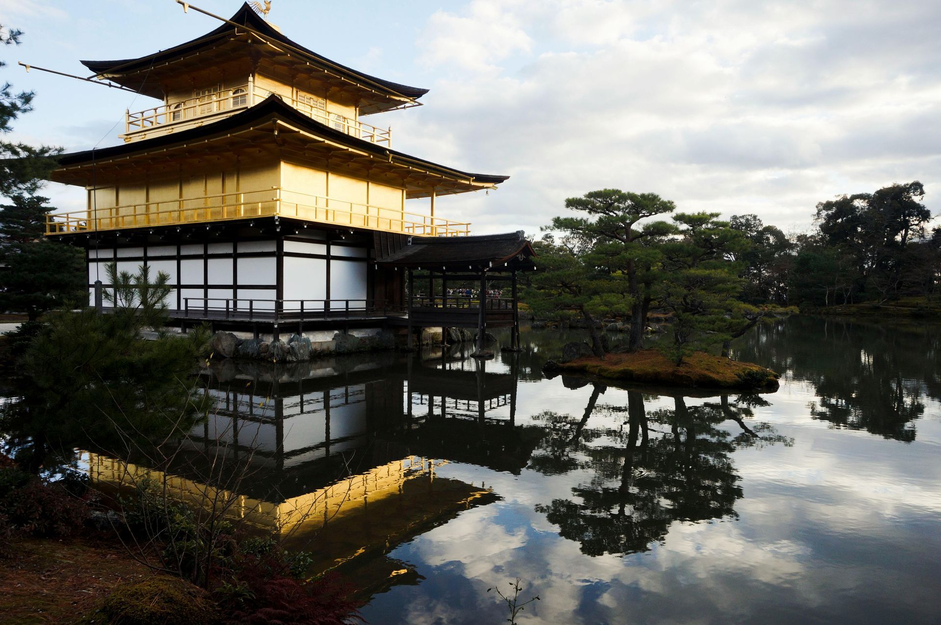 Golden Pavilion temple reflected in a pond; Kyoto, Japan. Gold building, dark roof, trees, and cloudy sky.