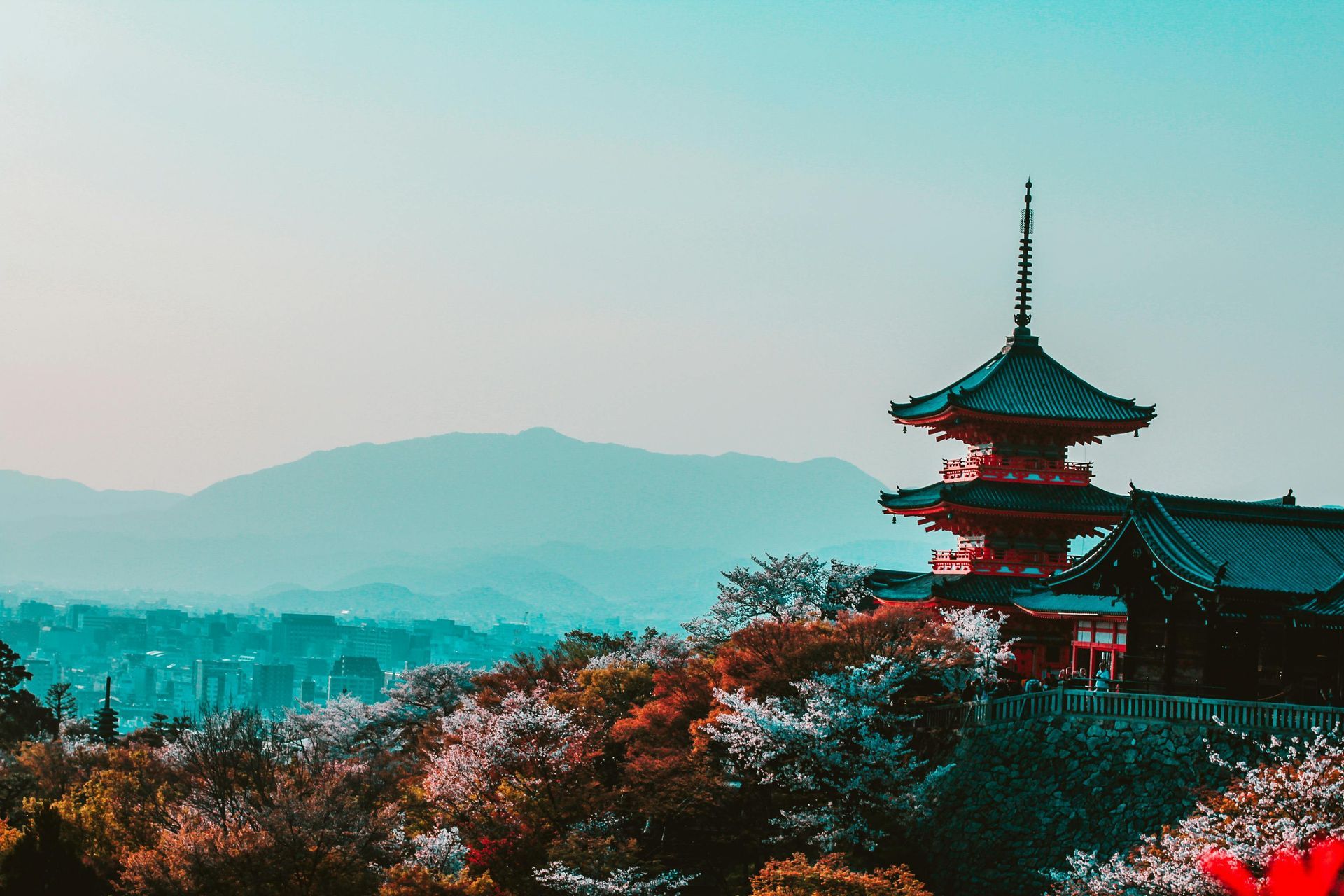 Red pagoda on a hillside, surrounded by blooming trees, mountains in the background, and a city skyline.