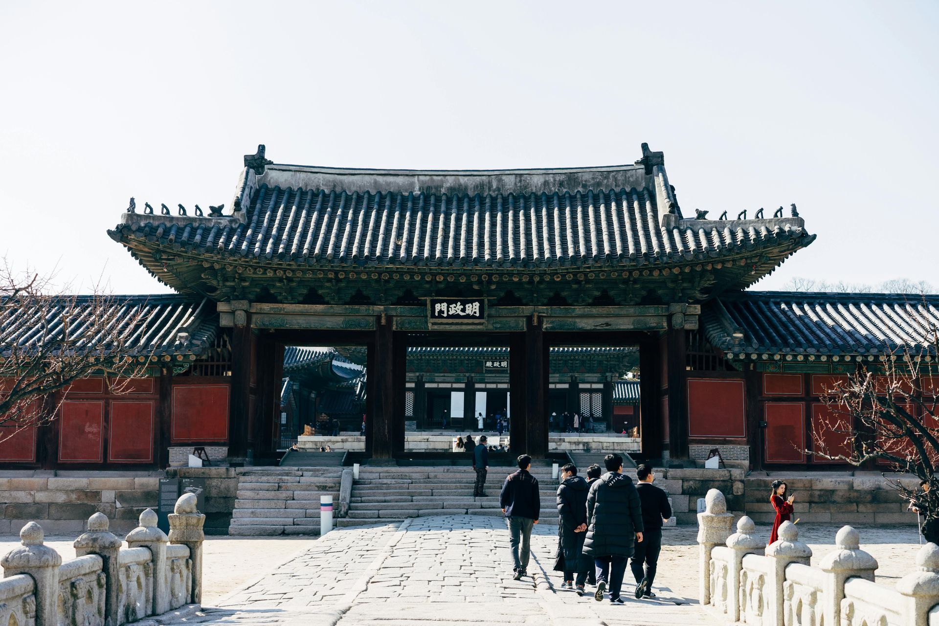 Traditional Korean gate with people walking through, red walls, stone bridge, gray tiled roof.