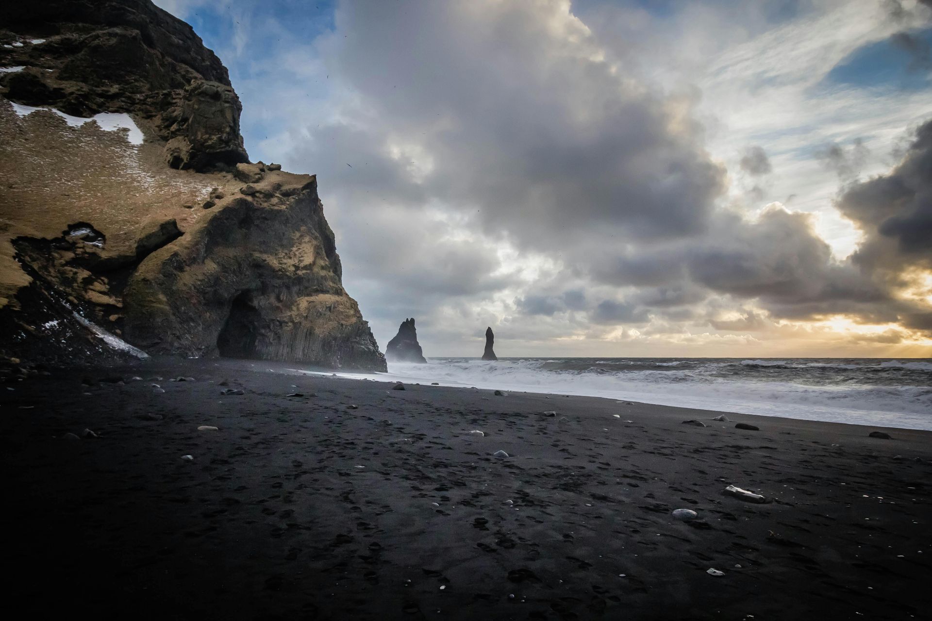 Black sand beach with dark cliffs and sea stacks under a dramatic, cloudy sky.