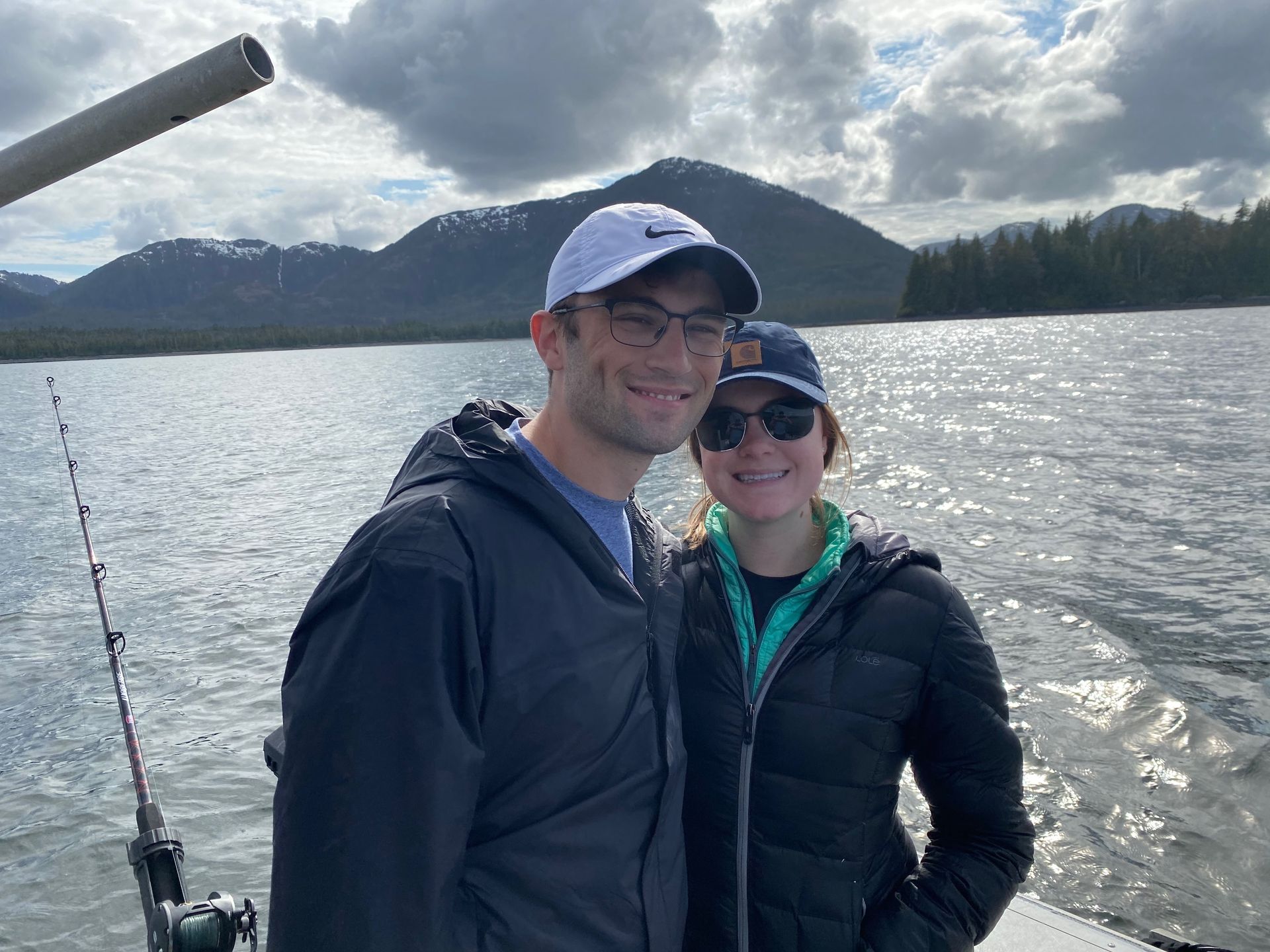 Woman on a boat practices yoga, arms raised, facing a lake and mountains at sunset.