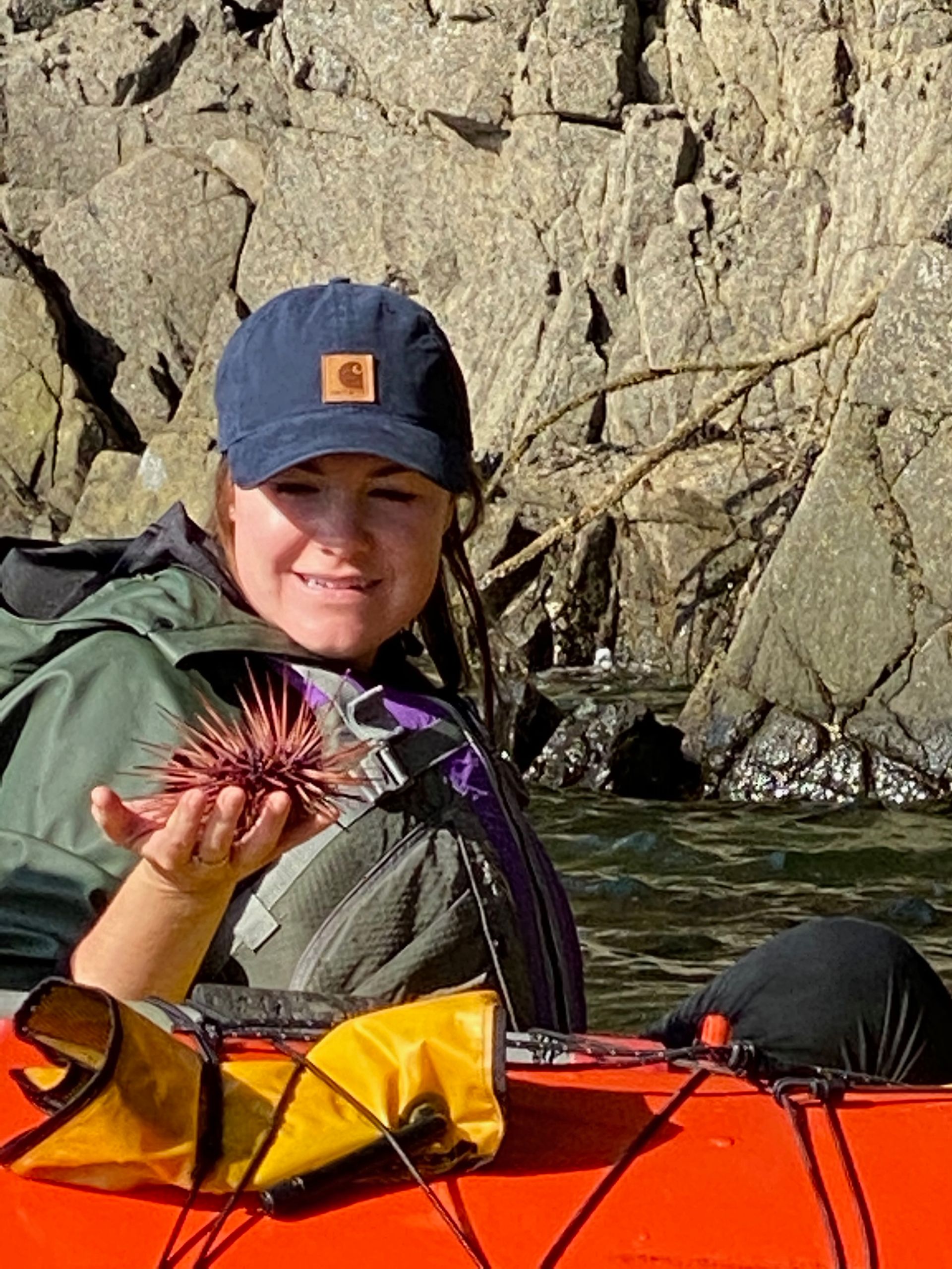 Woman in kayak holds a sea urchin. She wears a cap and life vest with rocky shore in background.