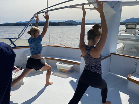 Two people doing yoga on a boat deck, arms raised, facing water and shoreline.