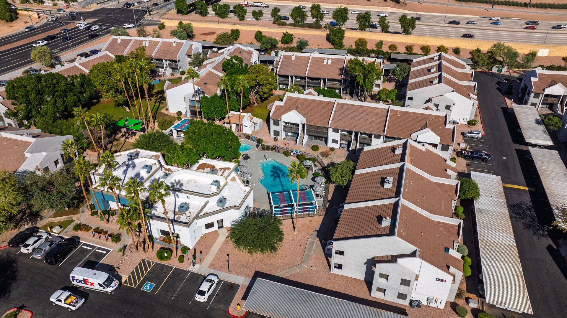 Aerial view of a complex with white buildings, brown roofs, pool, palm trees, parking lot.
