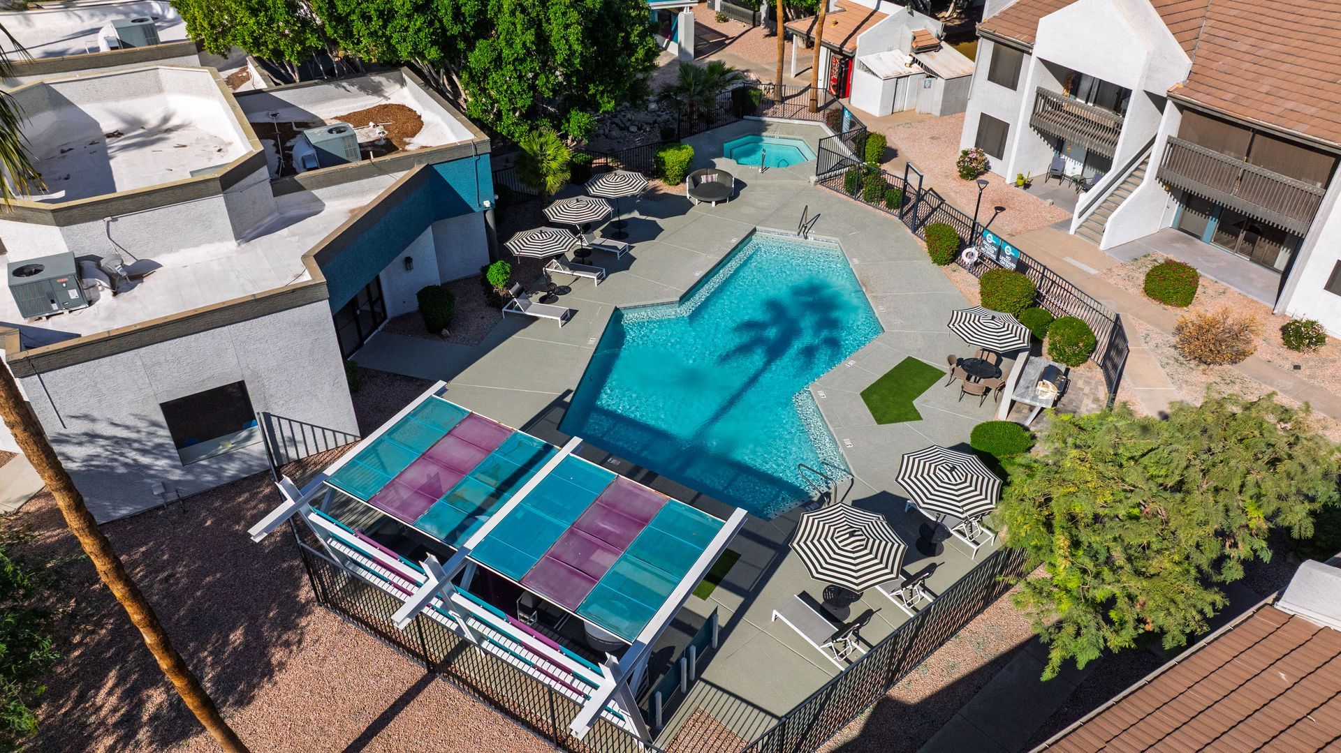Aerial view of a swimming pool with surrounding buildings, shaded seating, and palm tree shadow.