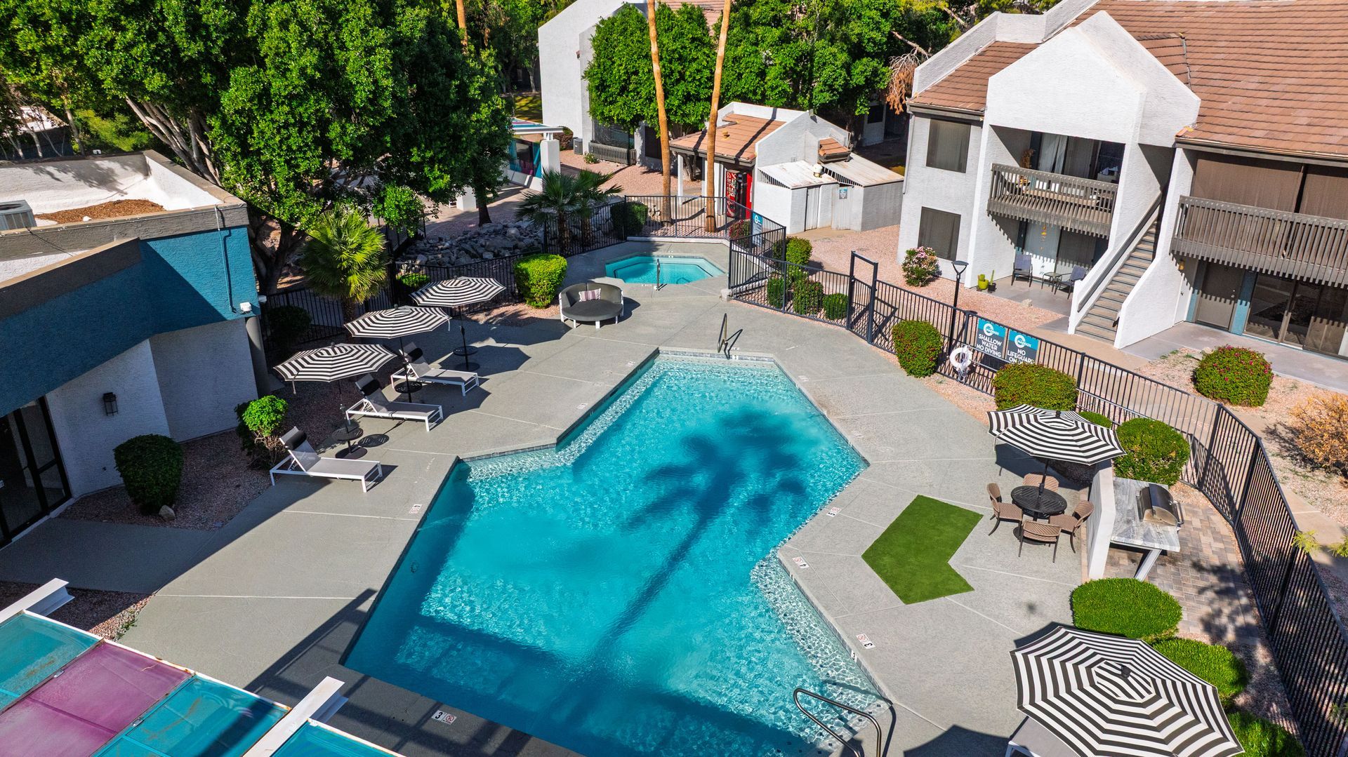 Pool area with lounge chairs, umbrellas, and buildings, trees.