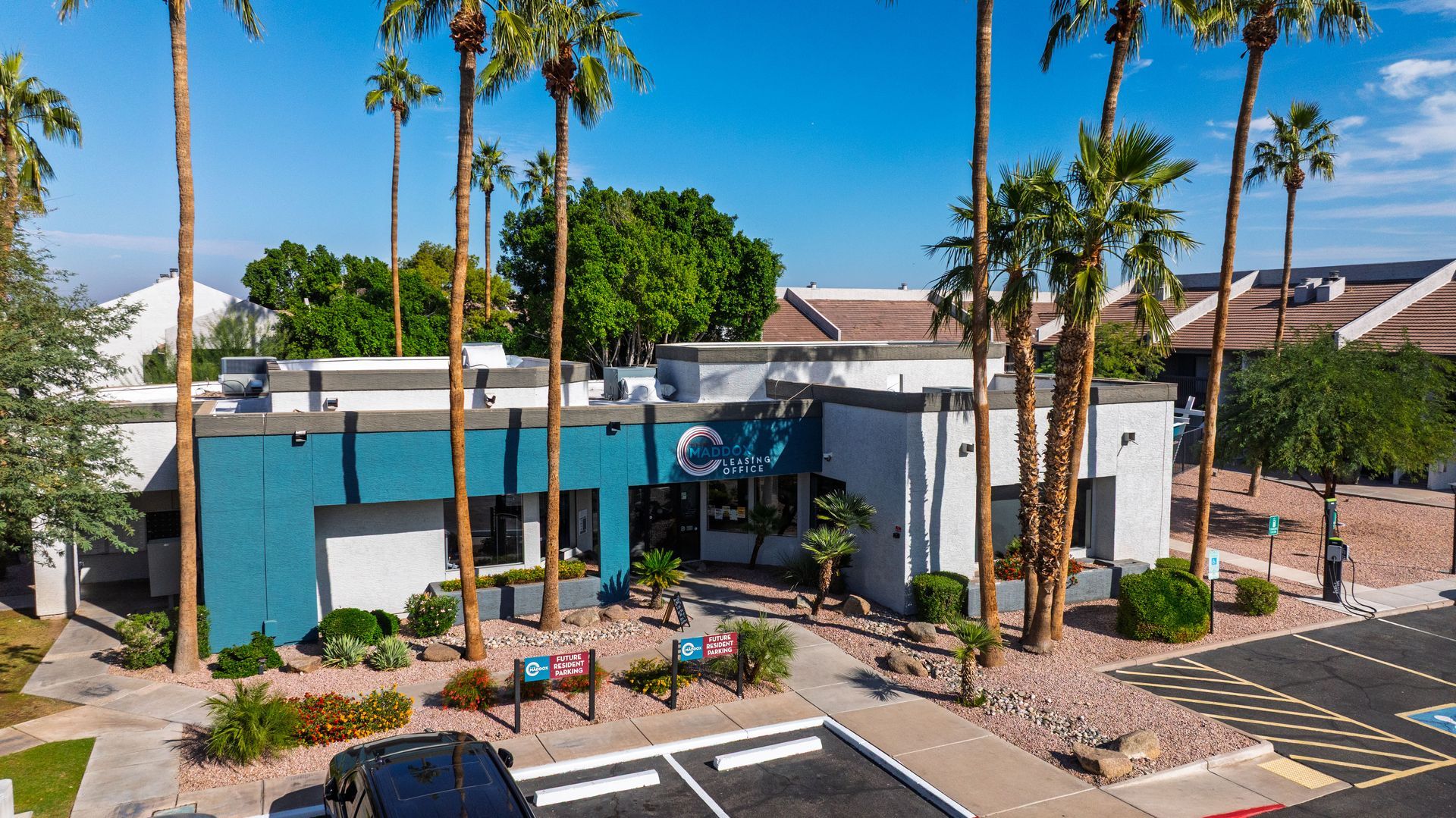 Modern building with teal facade, palm trees, and parking in sunny setting.