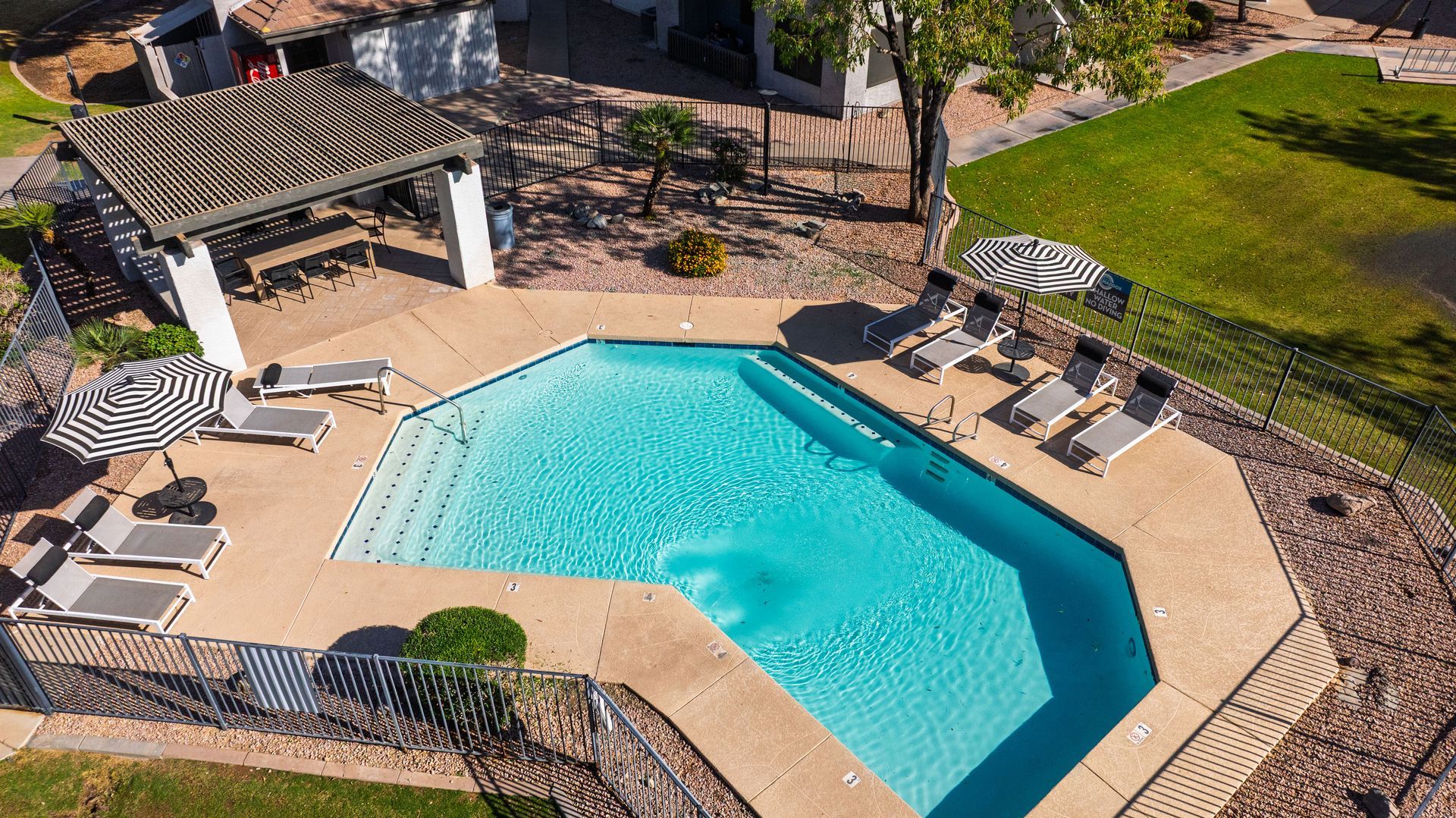 Swimming pool with lounge chairs, shaded pergola, and surrounding fence and grass.