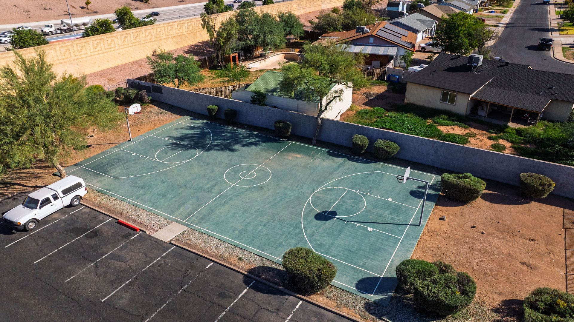 Aerial view of a green basketball court with white markings next to a parking lot and houses.