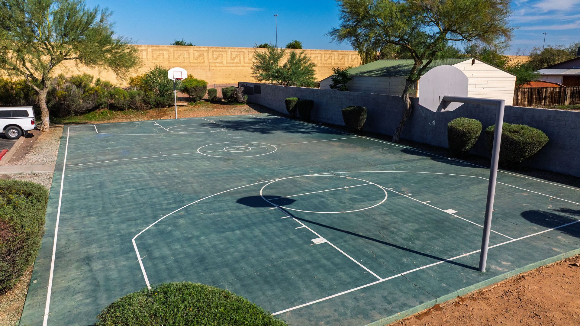 Outdoor basketball court with green surface, hoop, and backboard, surrounded by trees and a wall.
