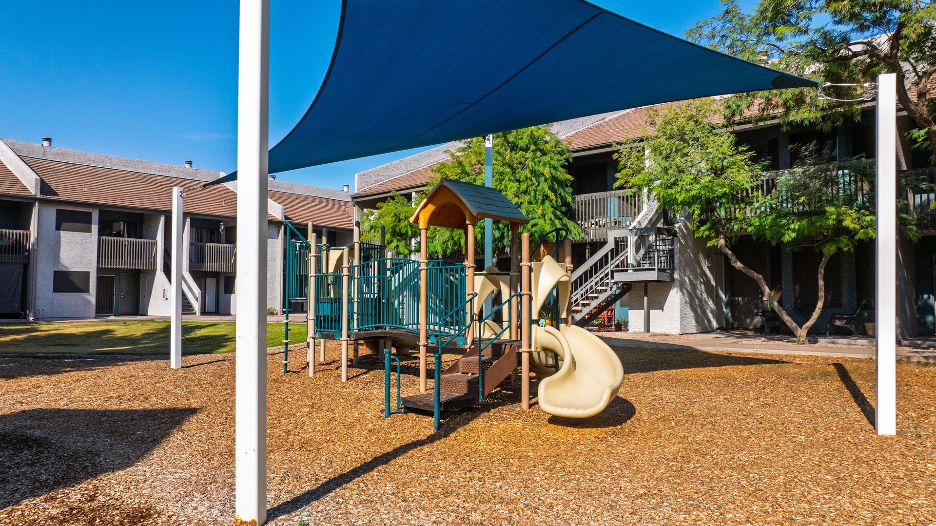 Playground with slide, under a blue shade, set in front of apartments.