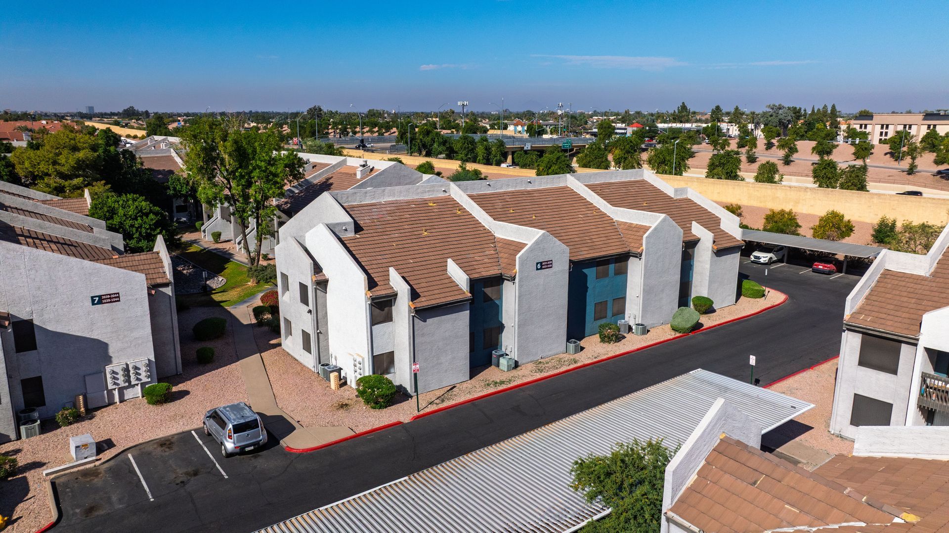 Aerial view of apartment complex with red-tiled roofs, parking, and surrounding landscape under a blue sky.