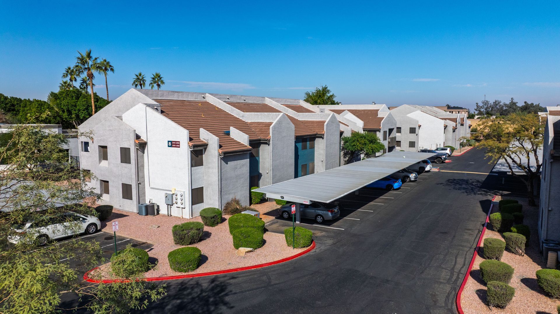 Apartment complex with a long covered parking area, palm trees, and blue sky.