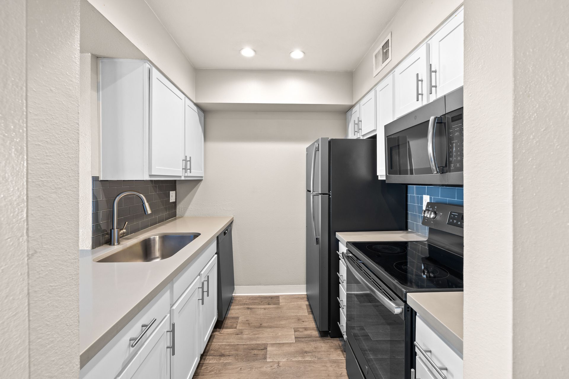 A galley kitchen with white cabinets, stainless steel sink, black appliances, and light wood flooring.