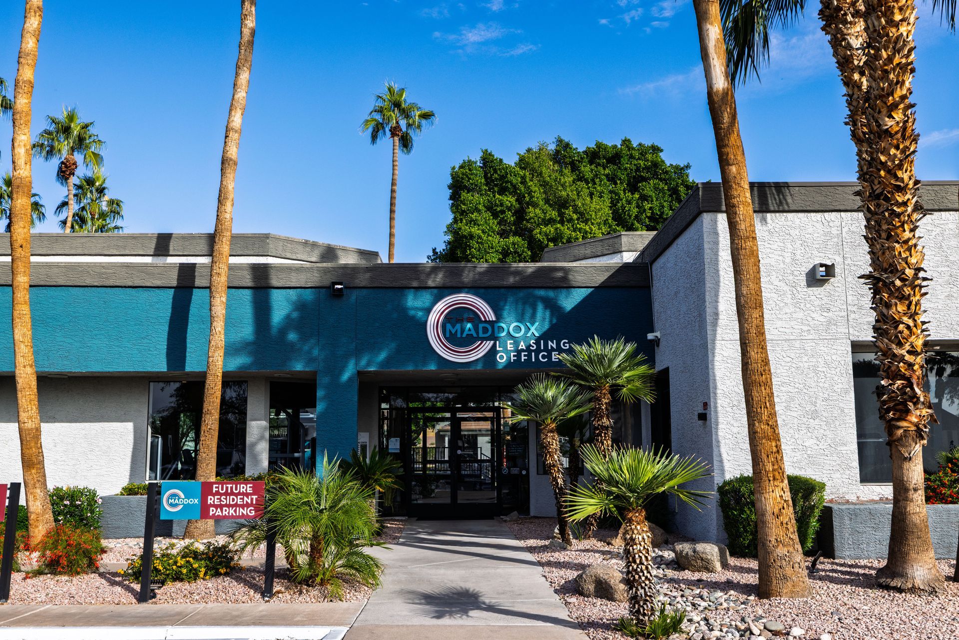Exterior of a teal and gray building with palm trees. Entrance marked by sign.