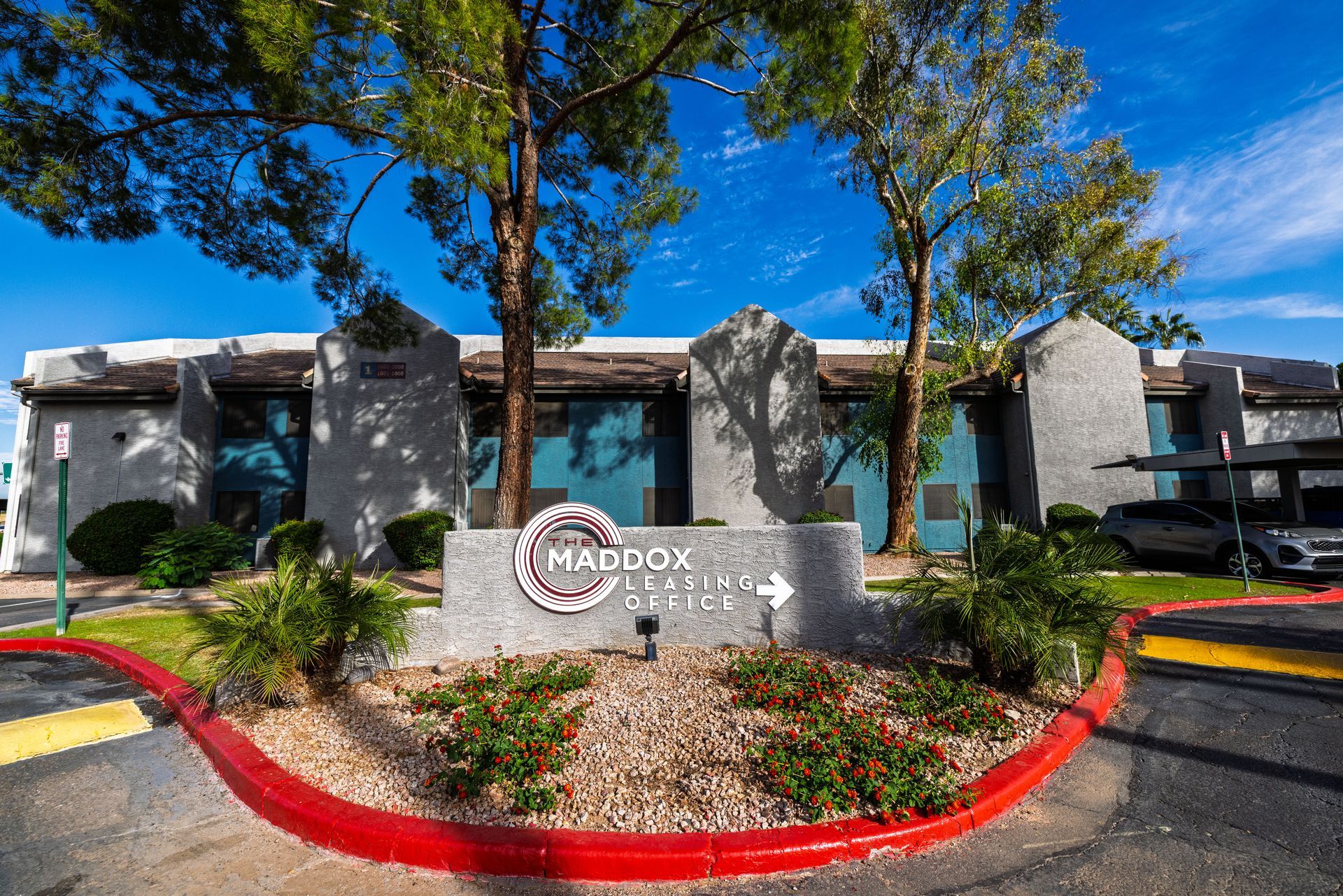 Sign for Maddox Apartments with an arrow, flanked by trees and bushes. Building in the background.