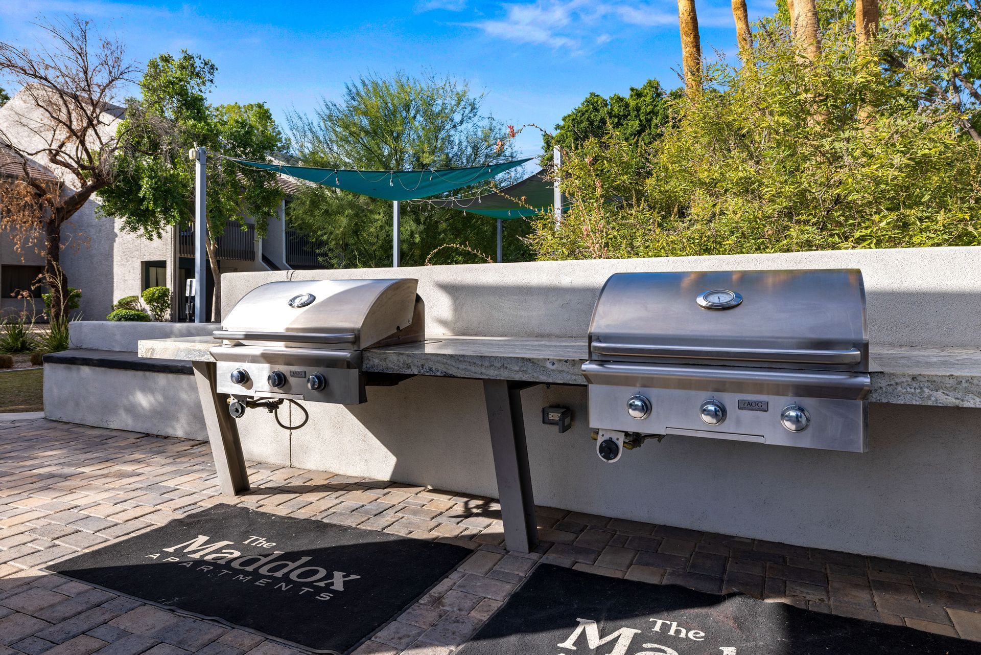Two stainless steel grills on a concrete counter, outdoor setting. Shade structure and landscaping in the background.