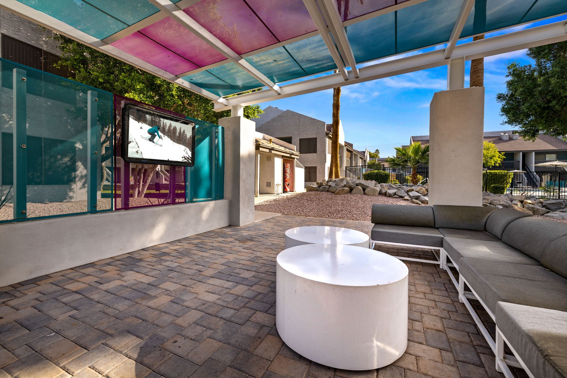 Outdoor lounge with a TV, couches, and white tables under a colorful canopy on a sunny day.